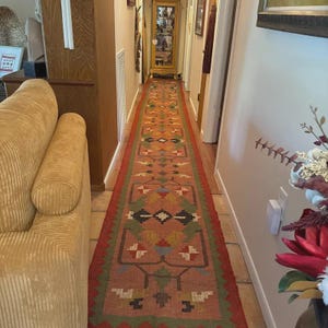 Pu&ograve; includere: Un tappeto da corridoio lungo e stretto con un motivo geometrico ripetitivo nei toni del terracotta, verde salvia, crema e marrone scuro. Il tappeto presenta un ampio bordo seghettato di un rosso intenso ed &egrave; posizionato sul pavimento.