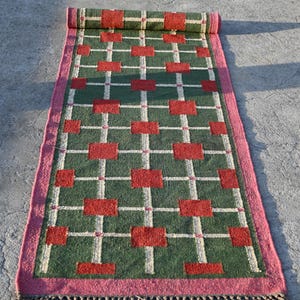 Könnte beinhalten: Ein rechteckiger Teppich mit grünem Hintergrund und einem sich wiederholenden Muster aus roten Quadraten und weißen Linien. Der Teppich hat einen rosa Rand und Fransen an einem Ende. Der Teppich ist oben aufgerollt.