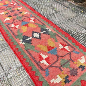 Pu&ograve; includere: Un lungo tappeto da corridoio rettangolare con uno sfondo rosso corallo vivace e un motivo floreale geometrico ripetuto nei toni del verde salvia, giallo senape, blu navy e crema. Il tappeto presenta un bordo rosso a zig-zag.