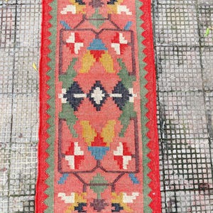 Pu&ograve; includere: Un tappeto passatoia rettangolare intrecciato con sfondo rosso corallo e un motivo geometrico nei toni del verde, giallo, blu, bianco e grigio scuro. Il tappeto presenta un bordo verde seghettato, un bordo esterno rosso e frange corte.