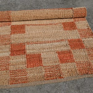 May include: A rolled-up, rectangular jute rug with a woven checkerboard pattern in natural and burnt orange tones. The rug is partially unrolled, displaying the pattern. The rug is made of a natural fiber.