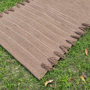 May include: A rectangular brown woven rug with vertical white stripes and tassels on the ends. The rug is lying on a bed of green grass. The rug is made of a natural fiber.