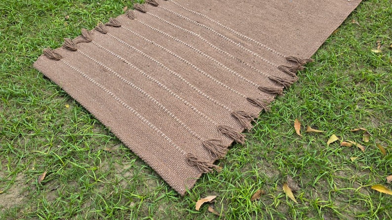 May include: A rectangular brown woven rug with vertical white stripes and tassels on the ends. The rug is lying on a bed of green grass. The rug is made of a natural fiber.