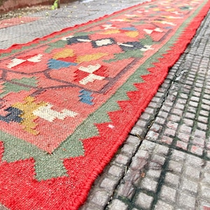 Pu&ograve; includere: Un lungo tappeto passatoia rettangolare con un colore di base rosso corallo vibrante. Il tappeto &egrave; decorato con un motivo geometrico ripetitivo nei toni del verde oliva, giallo senape, blu navy e bianco, con un bordo rosso a zig-zag.