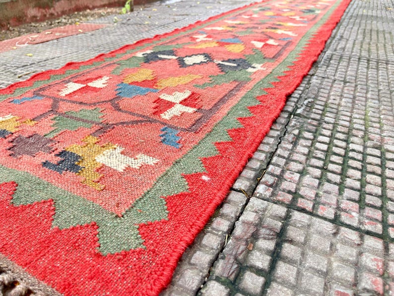 Pu&ograve; includere: Un lungo tappeto passatoia rettangolare con un colore di base rosso corallo vibrante. Il tappeto &egrave; decorato con un motivo geometrico ripetitivo nei toni del verde oliva, giallo senape, blu navy e bianco, con un bordo rosso a zig-zag.
