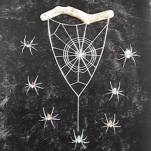 Peut inclure: Un fond noir avec une toile d'araignée en fil de fer blanc suspendue à un morceau de bois flotté. Il y a huit araignées argentées avec des perles colorées pour les yeux qui pendent de la toile.