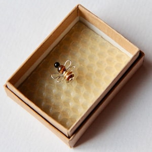 May include: A small, brown and black bee made of beads and wire is displayed in a cardboard box with a honeycomb pattern.