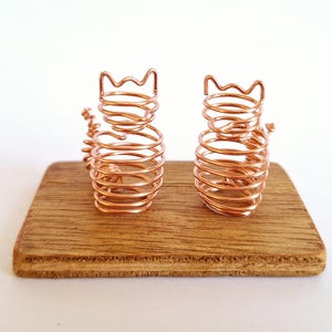 May include: Two copper wire cat sculptures with spiral bodies and crown-shaped heads sit on a small, rectangular wooden base. The cats are made of coiled copper wire, and the wooden base has a natural wood grain.