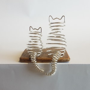 May include: Two silver wire sculptures of cats sitting side-by-side on a wooden base. The cats are made of coiled wire and have a whimsical, modern design.