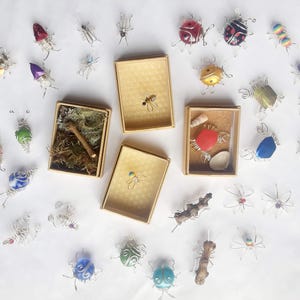 May include: A collection of handmade wire insect and bug sculptures with colorful glass beads. The sculptures are displayed on a white surface with small boxes and natural elements.