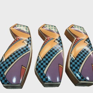 May include: Three abstract ceramic sculptures with a geometric design. The sculptures feature a combination of colours, including orange, purple, blue, and green, with a checkered pattern. The forms have a curved shape and are set against a white background.