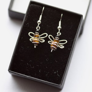 May include: A pair of bee-shaped earrings presented in a black box. The earrings have silver wire wings and a body made of amber-coloured beads and wire. Black beads form the head. The earrings hang from silver hooks.