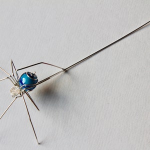 May include: A silver wire spider sculpture with a blue and white enamel bead for a body. The spider is suspended from a long silver wire with a loop at the end.
