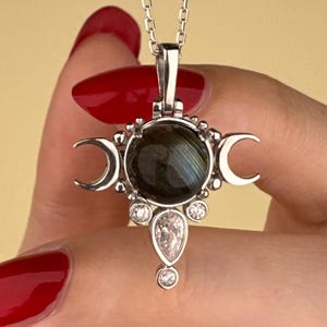 May include: Silver pendant necklace with a central dark gemstone, crescent moon accents, and clear crystal embellishments. The pendant hangs from a fine silver chain. The necklace is displayed against a pale yellow backdrop, creating a striking contrast. A mystical piece of jewellery.