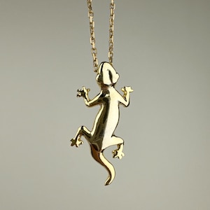Puede incluir: Un collar con colgante de lagarto dorado. El lagarto está detallado con extremidades extendidas y una cola curvada, suspendido de una delicada cadena. El colgante mide aproximadamente 5 cm de largo, y la cadena mide aproximadamente 46 cm.