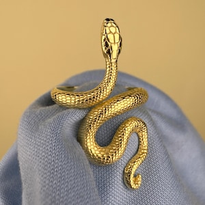 Könnte beinhalten: Ein goldfarbener Schlangenring mit strukturierter Oberfläche. Der Schlangenkopf ist erhoben, und der Körper windet sich. Der Ring wird auf einem hellblauen Stoff mit beigem Hintergrund präsentiert.