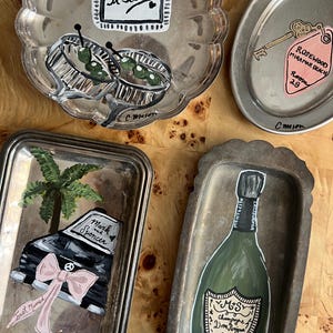 May include: A collection of hand-painted silver trays with various designs. One tray features the text "and so it bebenz", another depicts a car with a bow and the text "Just Married" and "Mark Spencer". Other trays show a key tag and a champagne bottle.