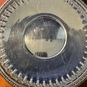 May include: A round silver-toned serving tray featuring a polished, reflective center surface with visible surface scratches. The wide, decorative border displays an intricate, repeating embossed pattern with a scalloped edge design.