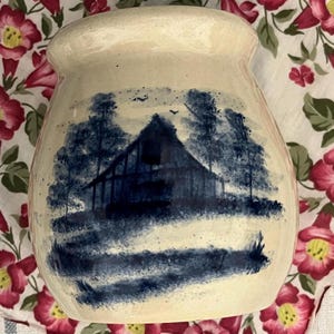 May include: Cream-colored ceramic jar with a blue and white barn scene. The jar features a wide opening and a slightly flared rim. The barn is depicted with trees and a field in a folk art style.