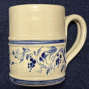 May include: A cylindrical ceramic mug with a speckled cream-colored finish. The mug features a prominent blue hand-painted floral and vine pattern encircling the lower section, accented by horizontal blue bands at the top and bottom edges.