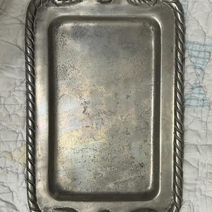 May include: A rectangular silver-toned metal tray featuring a rope-style border and decorative shell motifs at the top and bottom center, flanked by circular scroll patterns. The surface has a weathered, tarnished finish with visible texture.