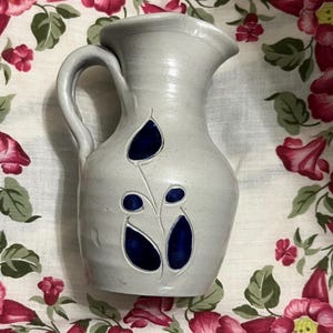May include: A gray ceramic pitcher with a handle and a spout. The pitcher is decorated with a blue floral design. The pitcher is sitting on a floral patterned fabric.