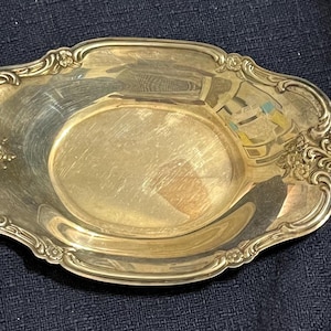 May include: An oval-shaped gold-colored metal tray with a polished, reflective surface. The rim features an ornate, raised border with intricate floral and scrollwork detailing along the edges, providing a decorative frame for the smooth center.