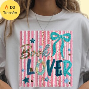 Peut inclure: Un t-shirt blanc avec un motif rectangulaire à rayures verticales roses et blanches. Le texte "Book Lover" est écrit en grandes lettres texturées, accompagné d'un grand nœud bleu sarcelle, de petites étoiles et de minuscules icônes de livres.
