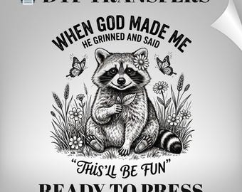 May include: DTF transfer design featuring a raccoon with a flower crown, holding a leaf, surrounded by butterflies and flowers. Text reads: "WHEN GOD MADE ME HE GRINNED AND SAID 'THIS'LL BE FUN' READY TO PRESS".