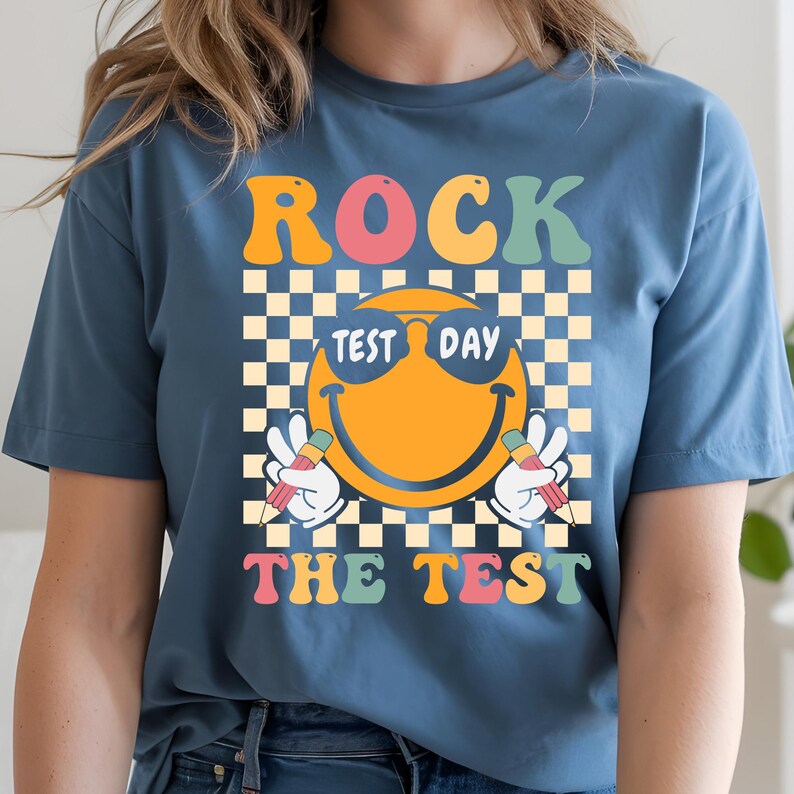 May include: A blue t-shirt with the words "ROCK THE TEST" in colorful, retro-style lettering. The design features a smiling emoji wearing sunglasses, holding pencils, and set against a checkered background. The shirt is made of a soft material.