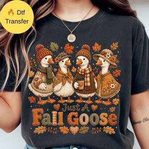 May include: A dark gray t-shirt featuring a graphic of four geese wearing autumn-themed clothing, including knit hats, scarves, and capes, surrounded by falling leaves. The text "Just A Fall Goose" is printed in large, textured, orange and brown letters.