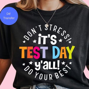 May include: Black t-shirt with a colorful graphic that reads "DON'T STRESS! IT'S TEST DAY y'all! DO YOUR BEST." The text is in a variety of colors, including orange, blue, and yellow. The shirt has a crew neck.