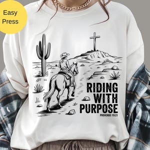 May include: White sweatshirt featuring a black and white illustration of a person riding a horse through a desert landscape towards a cross. The text "RIDING WITH PURPOSE" and "PROVERBS 19:21" are printed in black.