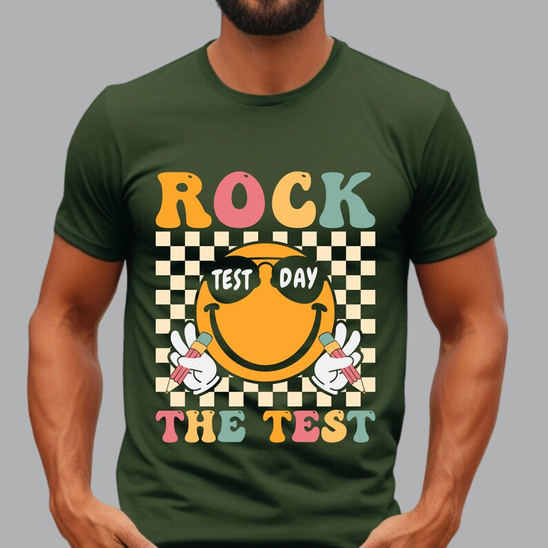 May include: Green t-shirt with a retro design. The graphic features the words "ROCK THE TEST" in orange, pink, and teal, with a smiling emoji wearing sunglasses and holding pencils. The background is a black and white checkered pattern.