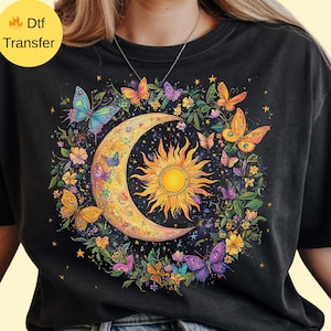 May include: Black t-shirt featuring a celestial design. The graphic includes a crescent moon, a radiant sun, and colorful butterflies. The design is surrounded by flowers and stars, creating a whimsical and artistic aesthetic.