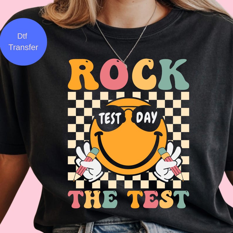 May include: Black t-shirt with a retro design featuring the words "ROCK THE TEST" in orange, pink, and green. A smiling emoji wearing sunglasses is in the center, with a checkered background. The emoji is holding pencils.