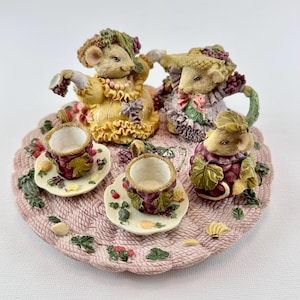 May include: A decorative figurine featuring four mice dressed in vintage clothing, gathered around a miniature tea set. The tea cups and saucers are decorated with grape and leaf motifs. The mice are posed on a pink, textured serving tray.