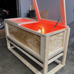 May include: A wooden incubator with a mesh-covered lid, open to reveal an orange-lit interior. The structure is supported by a wooden frame. The design suggests a handcrafted or DIY project, possibly for reptile or poultry incubation.
