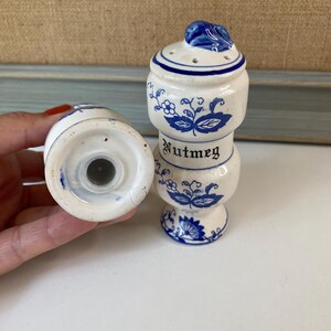 May include: White ceramic nutmeg shaker with blue floral designs and the word "Nutmeg" in black lettering. The shaker has a blue leaf-shaped top and small holes for dispensing. A second shaker is visible.