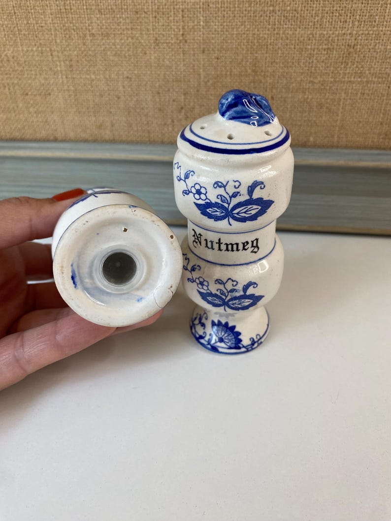 May include: White ceramic nutmeg shaker with blue floral designs and the word "Nutmeg" in black lettering. The shaker has a blue leaf-shaped top and small holes for dispensing. A second shaker is visible.