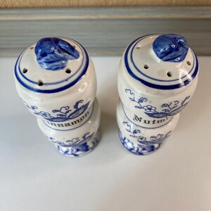 May include: Two ceramic spice shakers with blue and white floral designs. One shaker is labeled "Cinnamon" and the other "Nutmeg". Each shaker has a blue lid with a small handle and several holes for dispensing.