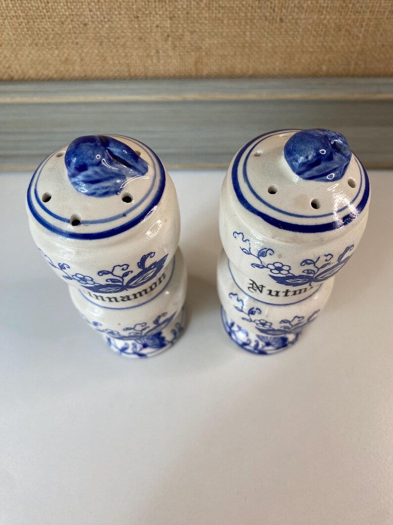 May include: Two ceramic spice shakers with blue and white floral designs. One shaker is labeled "Cinnamon" and the other "Nutmeg". Each shaker has a blue lid with a small handle and several holes for dispensing.