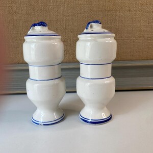May include: Two white ceramic salt and pepper shakers with blue trim. Each shaker has a bulbous body, a narrow waist, and a flared base. The lids are also white with blue accents and a small handle. The shakers are displayed on a white surface.