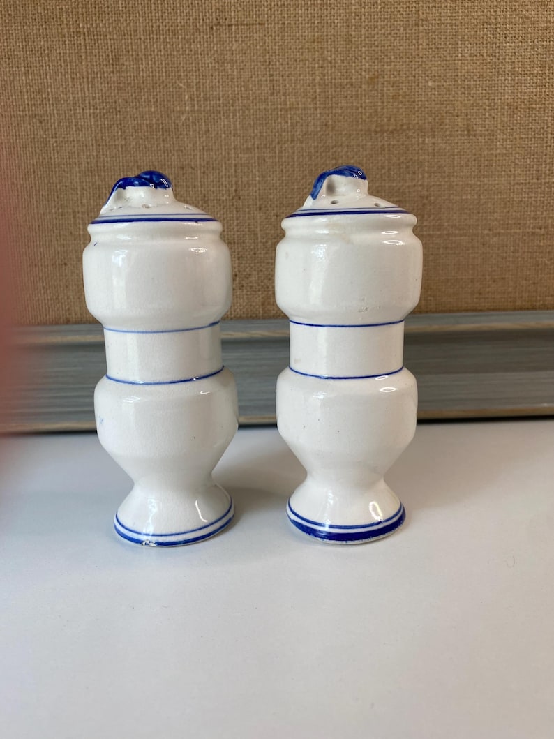 May include: Two white ceramic salt and pepper shakers with blue trim. Each shaker has a bulbous body, a narrow waist, and a flared base. The lids are also white with blue accents and a small handle. The shakers are displayed on a white surface.