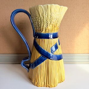 May include: A ceramic pitcher shaped like a bundle of yellow wheat stalks tied with a glossy blue ribbon. The pitcher features a tall, flared rim and a curved blue handle extending from the side, with textured details mimicking individual grain.