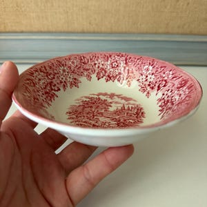 May include: A small, round ceramic bowl with a cream-colored base and a red transferware pattern. The interior features a detailed landscape scene at the center, while the rim is decorated with a dense floral motif of blossoms and leaves.