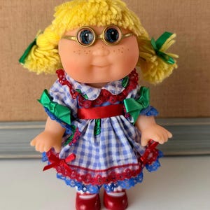 May include: A doll with yellow yarn hair styled in pigtails, wearing gold-rimmed round glasses. The doll has blue eyes and freckles, dressed in a blue and white gingham dress with red lace trim, a red ribbon waistband, and green ribbon accents.