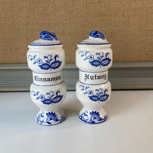 May include: Two white ceramic spice jars with blue floral designs and lids. One jar is labeled "Cinnamon" and the other "Nutmeg". The jars are stacked with a bulbous base and a small handle on the lid.