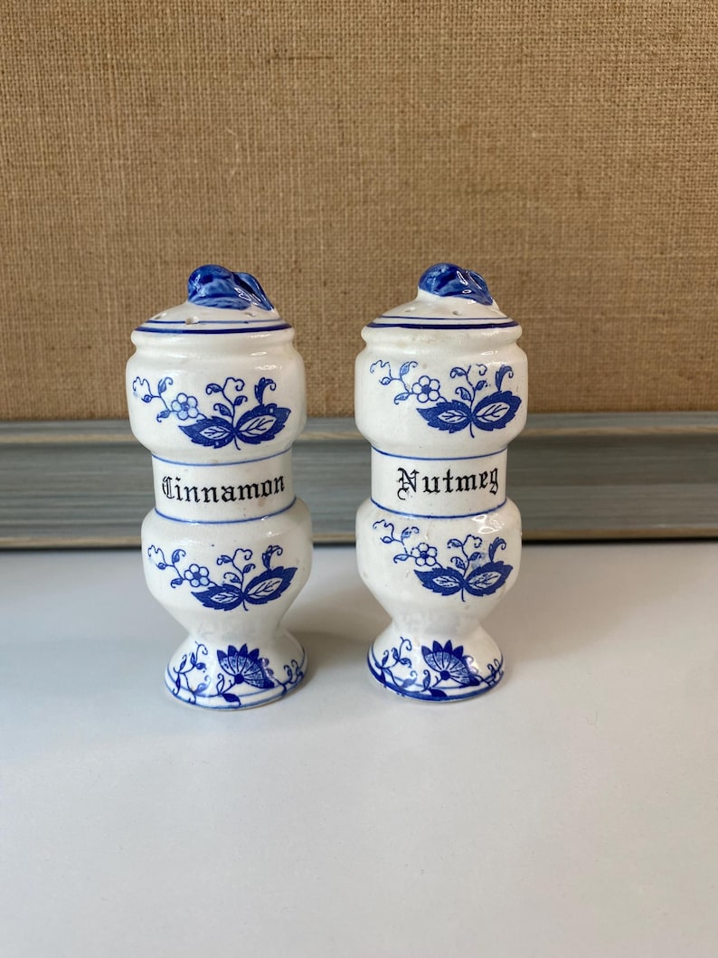 May include: Two white ceramic spice jars with blue floral designs and lids. One jar is labeled "Cinnamon" and the other "Nutmeg". The jars are stacked with a bulbous base and a small handle on the lid.