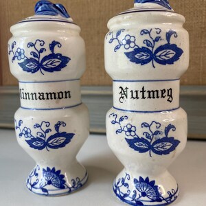 May include: Two white ceramic spice jars with blue floral designs and lids. One jar reads "Cinnamon" and the other reads "Nutmeg" in black lettering. The jars have a bulbous shape and are approximately 4 inches tall.